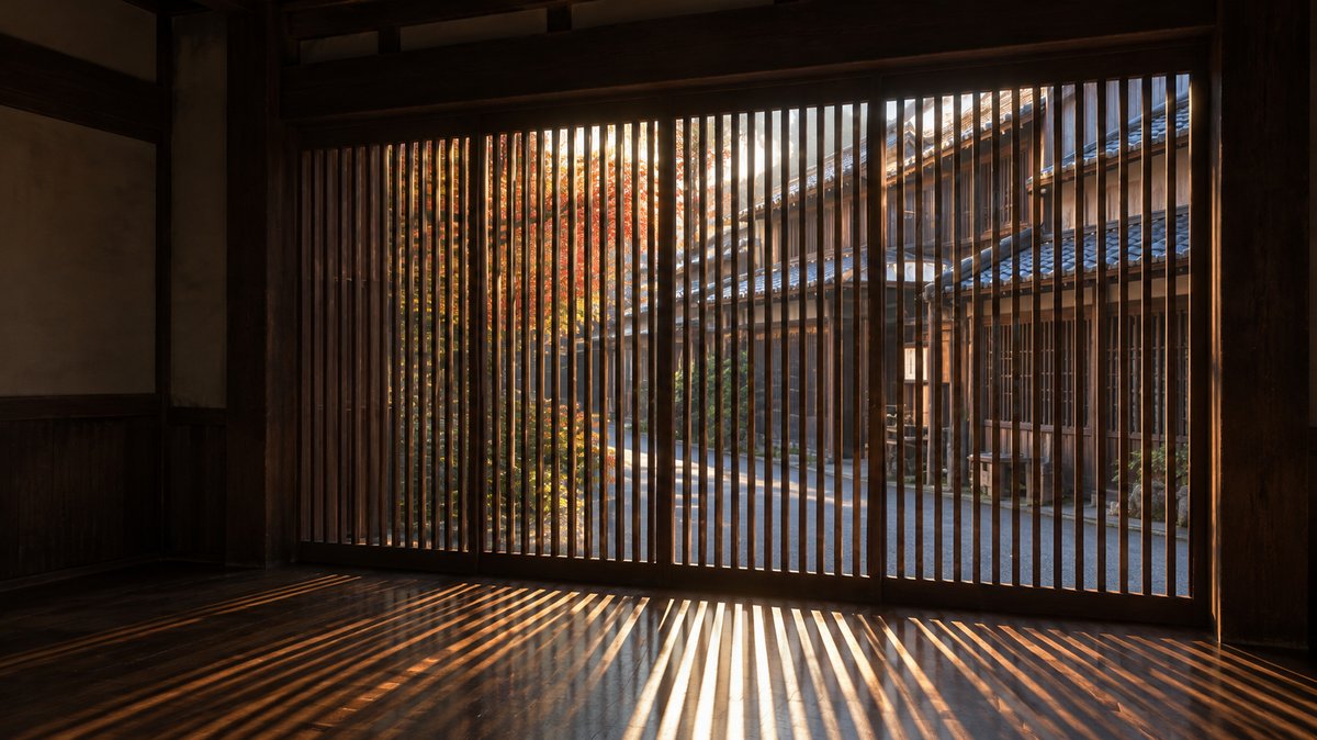 From inside a dim wooden townhouse, a large window of vertical wooden slats rises from the floor nearly to the ceiling. Through the narrow gaps, a bright autumn lane is visible, with a row of old wooden townhouses under tiled roofs and a tree in red and yellow leaves. Low sunlight passes through the slats and lays many thin golden stripes of light across the dark, polished wooden floor, while the walls and pillars of the room stay in deep shadow.